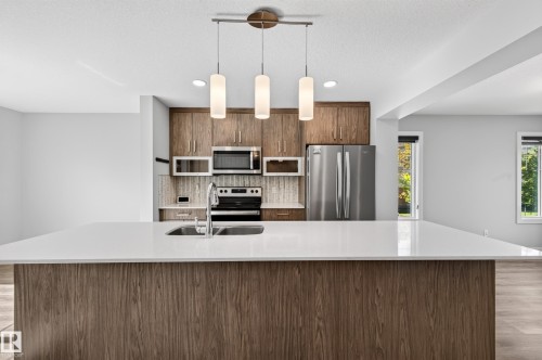 Kitchen featuring a large island with a white countertop, wood-finish cabinetry, stainless steel appliances, and pendant lighting - #44 3025 151 Av Nw, Edmonton, AB 