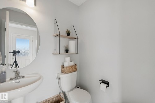 Pedestal sink with chrome faucet, circular wall mirror, wall-mounted light fixture, and a two-tier wall shelf with wood-finish shelves and black metal supports - 8535 Ellis Link Link Nw, Edmonton, AB 
