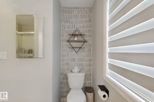 White toilet with a patterned accent wall, geometric wall-mounted shelving, and a horizontal striped window blind - 8535 Ellis Link Link Nw, Edmonton, AB 
