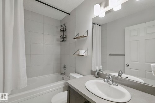 White subway-tiled shower surround with a built-in tub, a single-sink vanity with a light gray countertop, and a vanity mirror with overhead lighting - 8535 Ellis Link Link Nw, Edmonton, AB 