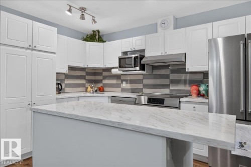 White shaker-style cabinetry, stainless steel appliances, patterned tile backsplash, track lighting, and a kitchen island with a light-colored countertop - #125 4610 50 Ave, Stony Plain, AB 