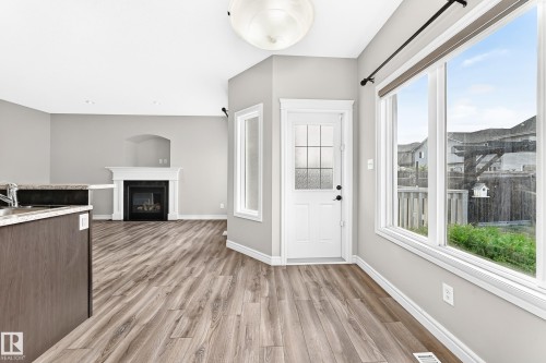 Wood-finish flooring throughout, white framed gas fireplace with mantel, white paneled door with window pane inserts, large window with white trim, and a built-in kitchen island with stone-finish countertop - Edmonton, AB 