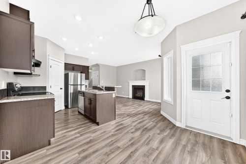 Wood-finish flooring throughout, kitchen with dark wood cabinetry and stainless steel appliances, central island with seating, recessed lighting, and a fireplace with a mantel - Edmonton, AB 
