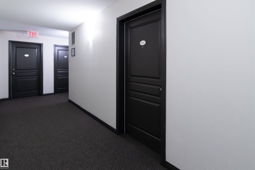 Corridor featuring dark paneled entry doors, textured white walls, recessed ceiling lighting, and dark carpet flooring - #214 1188 Hyndman Rd Nw, Edmonton, AB 
