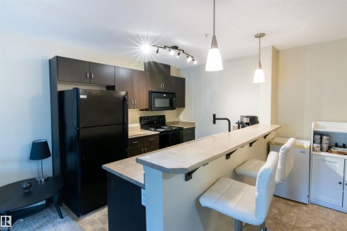 Kitchen featuring a breakfast bar with two pendant lights, dark wood-finish cabinetry, black appliances including a refrigerator and range, and track lighting - #214 1188 Hyndman Rd Nw, Edmonton, AB 