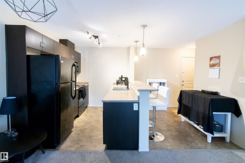 Modern kitchen featuring dark cabinetry, black appliances, light-toned countertops, and a breakfast bar with pendant lighting - #214 1188 Hyndman Rd Nw, Edmonton, AB 