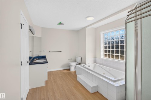 Bathroom featuring a built-in jetted tub, glass block window, wood-finish flooring, white vanity with an integrated sink, and a separate shower enclosure - 4409 150 Avenue Nw, Edmonton, AB 