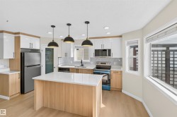 Modern kitchen featuring a central island with a light-toned countertop, wood-finish cabinetry, stainless steel appliances, and three pendant lights - 