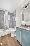 Wood-finish flooring, full bathtub with shower, gray tile surround, light blue vanity with frosted glass doors, and a back-lit mirror - 