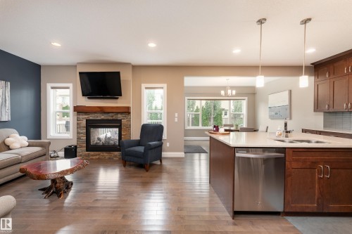 Spacious living area featuring wood-finish flooring, a stone-faced fireplace with a wood mantel, an open-concept kitchen with a large island, stainless steel appliances, and pendant lighting - 6613 31 Avenue, Beaumont, AB 