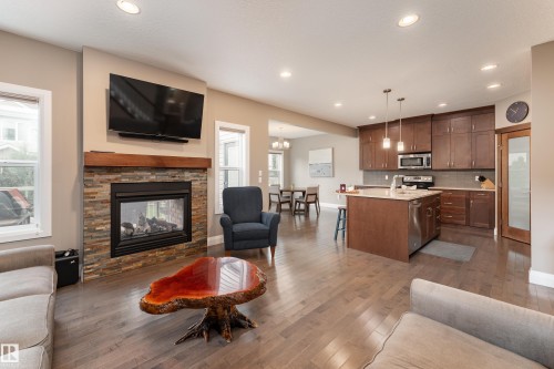 Stacked stone fireplace with a wood mantel, wood-finish flooring, recessed lighting, and a kitchen featuring wood cabinetry and an island with pendant lighting - 6613 31 Avenue, Beaumont, AB 