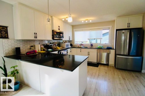 White shaker-style cabinetry with dark hardware, black countertops, hexagonal tile backsplash, stainless steel appliances, and wood-finish flooring - 17440 77 Street Nw, Edmonton, AB 