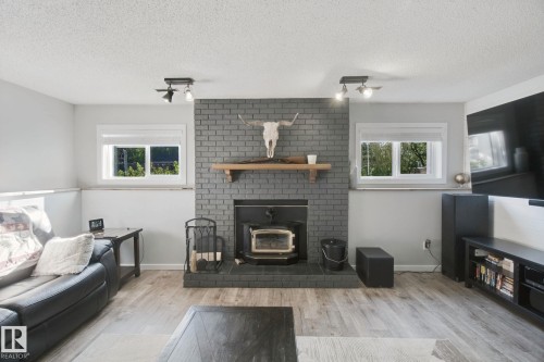 Brick fireplace with wood-burning stove, wood-finish flooring, recessed windows, and track lighting fixtures - 38 Menalta Place, Cardiff, AB 