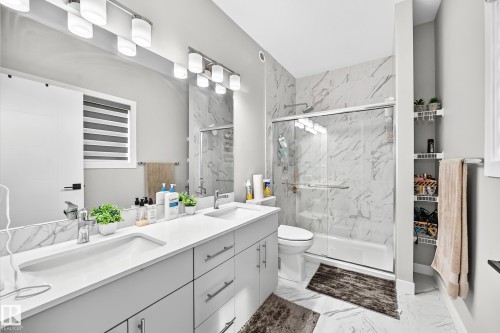 Dual vanity with undermount sinks and chrome faucets, full-width mirror, white cabinetry, shower-over-tub with sliding glass door, and marbled wall tile - 2415 194 Street Nw, Edmonton, AB 