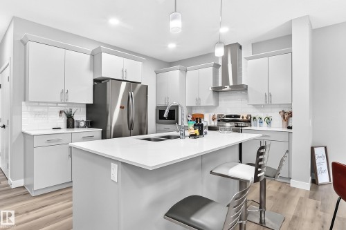 Kitchen featuring light-toned cabinetry, a central island with a sink, stainless steel appliances, a tile backsplash, and wood-finish flooring - 2415 194 Street Nw, Edmonton, AB 