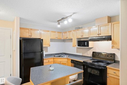Kitchen with light wood-finish cabinetry, dark countertops, black appliances, and a peninsula with seating - #203 245 Edwards Dr Sw, Edmonton, AB 