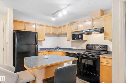 Kitchen featuring wood-finish cabinetry, black appliances, a double basin sink, and an integrated breakfast bar - #203 245 Edwards Dr Sw, Edmonton, AB 