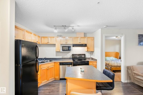 Kitchen featuring wood cabinetry, black appliances, a track lighting fixture, and an island with a breakfast bar - #203 245 Edwards Dr Sw, Edmonton, AB 