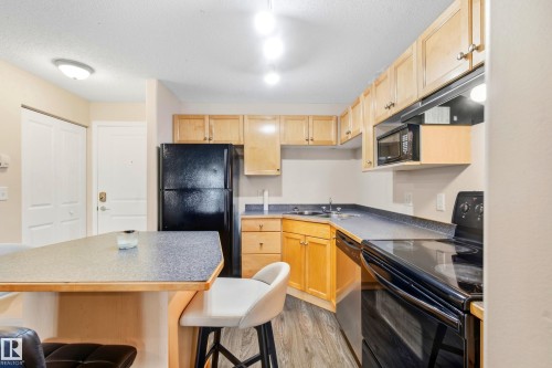 Kitchen featuring wood-finish flooring, light wood cabinetry, black appliances, and a breakfast bar - #203 245 Edwards Dr Sw, Edmonton, AB 