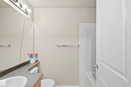 Built-in vanity with a white basin and wood-finish cabinetry, full-width mirror, and a white tiled shower-tub combination - #203 245 Edwards Dr Sw, Edmonton, AB 