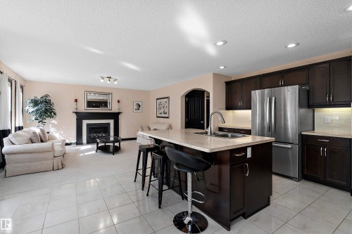 Kitchen island with seating, stainless steel refrigerator, dark wood-finish cabinetry, tile flooring, and a fireplace with mantel - 11515 16 Avenue Sw, Edmonton, AB 