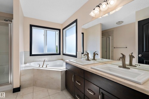 Corner soaking tub with tiled surround, dual windows with black trim, double vanity with integrated sinks, brushed nickel faucets, and a glass-enclosed shower - 11515 16 Avenue Sw, Edmonton, AB 
