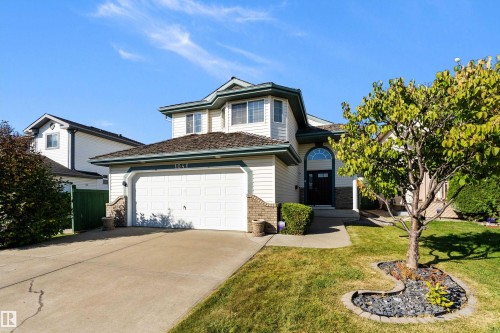 Two-story residence featuring light-toned siding, a two-car garage with a concrete driveway, a front lawn, and a mature tree with a stone-edged garden bed - 1046 Blackburn Close Sw, Edmonton, AB 