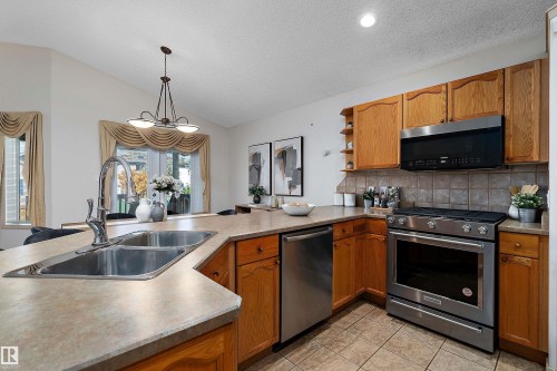 Kitchen featuring wood cabinetry, stainless steel appliances, a double basin sink, tile flooring, and vaulted ceilings - 1046 Blackburn Close Sw, Edmonton, AB 