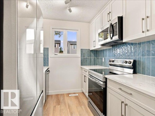 Modern kitchen featuring stainless steel appliances, white cabinetry with hardware, light-toned wood-finish flooring, and a blue tile backsplash - 18325 66 Avenue Nw, Edmonton, AB 