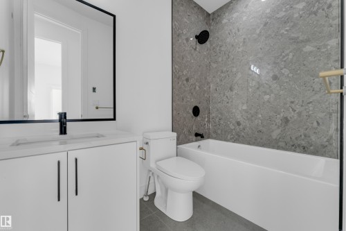 White vanity with integrated sink and matte black faucet, framed mirror, built-in bathtub, and gray stone-finish wall tiling - 512 Robinson Boulevard, Leduc, AB 