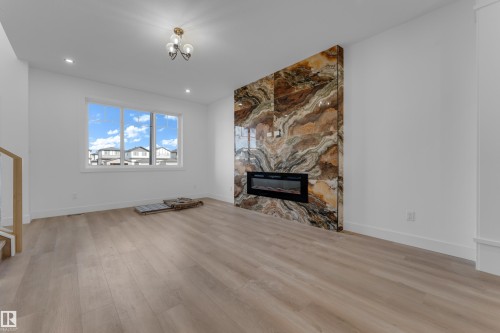 Spacious room featuring wood-finish flooring, a contemporary electric fireplace with a large stone surround, a prominent window with white trim, and recessed ceiling lighting - 512 Robinson Boulevard, Leduc, AB 
