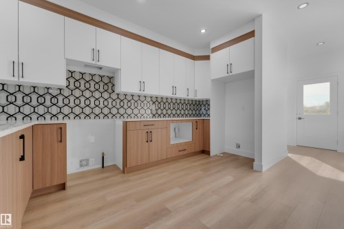 Two-tone cabinetry with matte black hardware, geometric tile backsplash, recessed lighting, wood-finish flooring, and a glass-paneled door - 512 Robinson Boulevard, Leduc, AB 