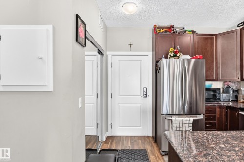 Stainless steel refrigerator, dark wood-finish cabinetry, granite-style countertop, wood-finish flooring, and a flush-mount ceiling light fixture - #404 12804 140 Av Nw, Edmonton, AB 