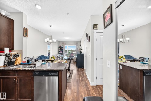 Kitchen featuring dark wood-finish cabinetry, stainless steel dishwasher, and a granite-style countertop island - #404 12804 140 Av Nw, Edmonton, AB 