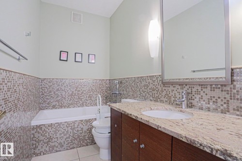 Bathroom featuring a built-in bathtub, mosaic tile surround, vanity with wood-finish cabinetry, stone countertop, and a large framed mirror - 995 Ogilvie Boulevard Nw, Edmonton, AB 