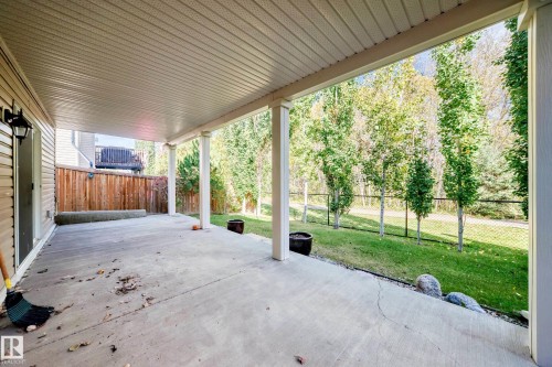 Covered concrete patio with white support columns, overlooking a grassy area with mature trees and a black chain-link fence - 21 Silverstone Drive, Stony Plain, AB 