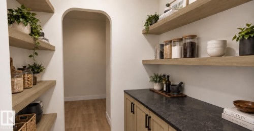 Integrated pantry shelving with wood-finish floating shelves, dark stone-look countertop, light wood-finish cabinetry with black hardware, and an arched doorway - 28 Brickyard View, Stony Plain, AB 