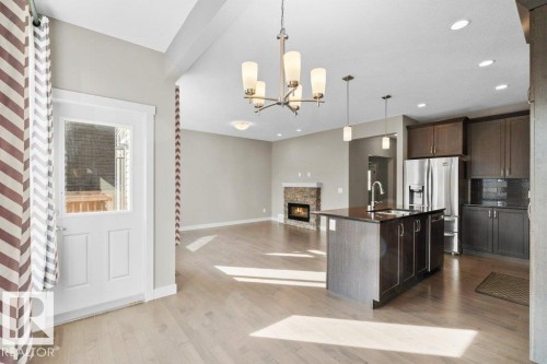 Open-concept interior featuring wood-finish flooring, a kitchen island with sink and pendant lighting, stainless steel refrigerator, dark wood-finish cabinetry, and a stone-clad fireplace - 1523 168 Street Sw, Edmonton, AB 
