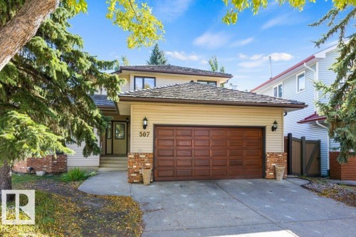 Front exterior featuring light-toned siding, a two-car garage with a wood-look door, brick accents, and a concrete driveway - 507 Burton Close Nw, Edmonton, AB 