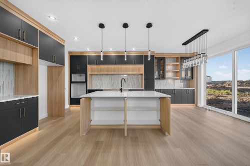 Kitchen featuring two-tone cabinetry with contrasting wood-finish and dark panels, a central island with a white countertop and integrated sink, stainless steel built-in appliances, pendant lighting, and wood-finish flooring - 13024 186 Avene Nw, Edmonton, AB 