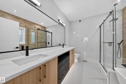 Dual vanity with integrated sinks and black matte faucets, expansive mirror with task lighting, wood-finish and dark cabinetry, large format wall and floor tiling, and a frameless glass shower enclosure - 
