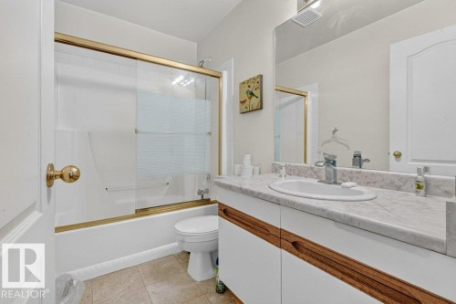 Bathroom featuring a white bathtub with gold-finish sliding glass doors, a vanity with a white countertop and integrated sink, a large mirror, and tiled flooring - Edmonton, AB 