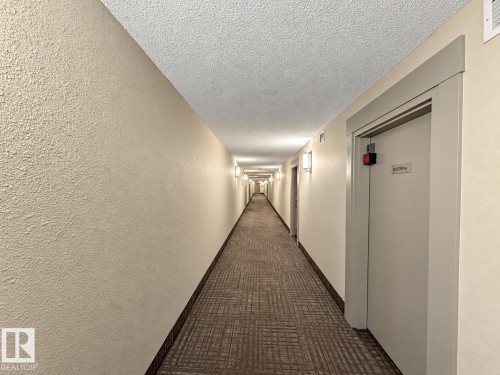 Hallway with textured neutral walls, carpeted flooring, and wall-mounted lighting fixtures - #116 4008 Savaryn Dr Sw Sw, Edmonton, AB 