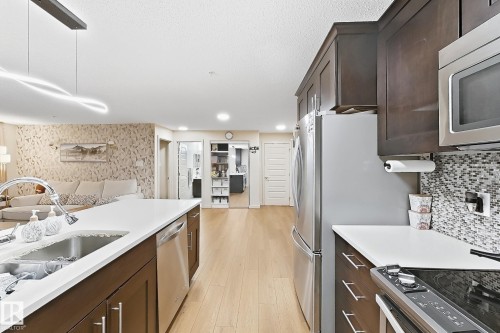 Kitchen featuring stainless steel appliances, dark wood-finish cabinetry, light-toned countertops, a mosaic tile backsplash, and light wood-finish flooring - #116 4008 Savaryn Dr Sw Sw, Edmonton, AB 