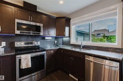 Kitchen featuring dark wood cabinetry, stainless steel appliances, speckled stone countertops, a tile backsplash, and a large window - 