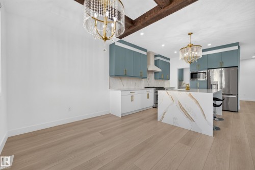 Modern kitchen featuring light wood-finish flooring, exposed ceiling beams, a waterfall island with a patterned surface, teal upper cabinetry, and stainless steel appliances - 11010 174A Avenue Nw, Edmonton, AB 