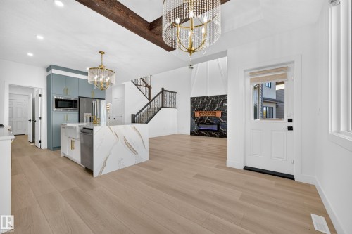 Open-concept living space with light wood-finish flooring, a kitchen island featuring a waterfall countertop, a black marble-style fireplace, and a striking staircase with dark balusters - 11010 174A Avenue Nw, Edmonton, AB 