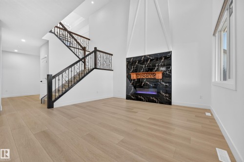 Expansive room featuring light wood-finish flooring, a contemporary fireplace with a black polished surround and carved wood mantel, and a two-story staircase with dark wood and wrought iron railings - 11010 174A Avenue Nw, Edmonton, AB 