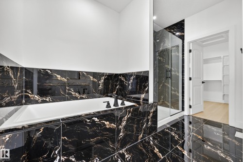 Deep soaking tub with matte black fixtures, full-height black and gold-veined tile, glass-enclosed shower, and built-in closet shelving - 11010 174A Avenue Nw, Edmonton, AB 