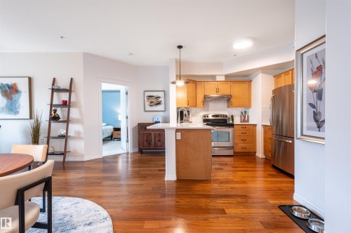Hardwood-finish flooring extending through the kitchen and living area, featuring light wood cabinetry, stainless steel appliances, a white tile backsplash, and a white kitchen island - #230 45 Inglewood Dr, St. Albert, AB 